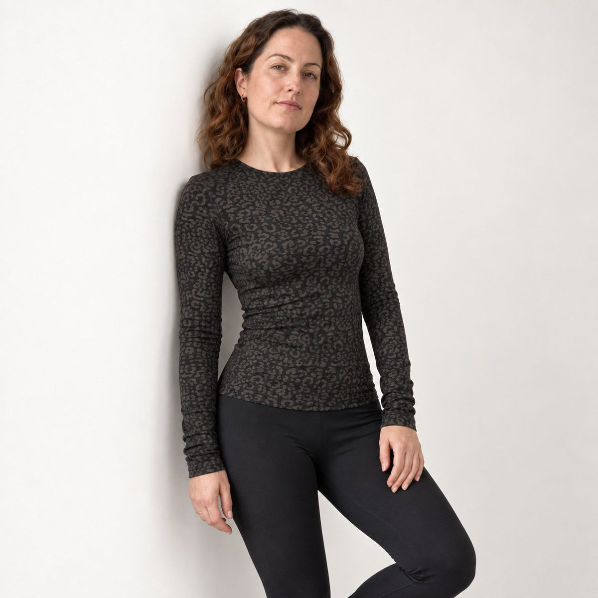 A woman wearing a fitted black leopard long sleeve top with a dark brown and black leopard print, paired with black leggings, posing against a plain off-white wall.