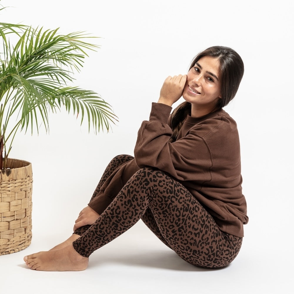 Woman wearing a brown sweatshirt and leopard print pants sitting next to a potted plant on a white background