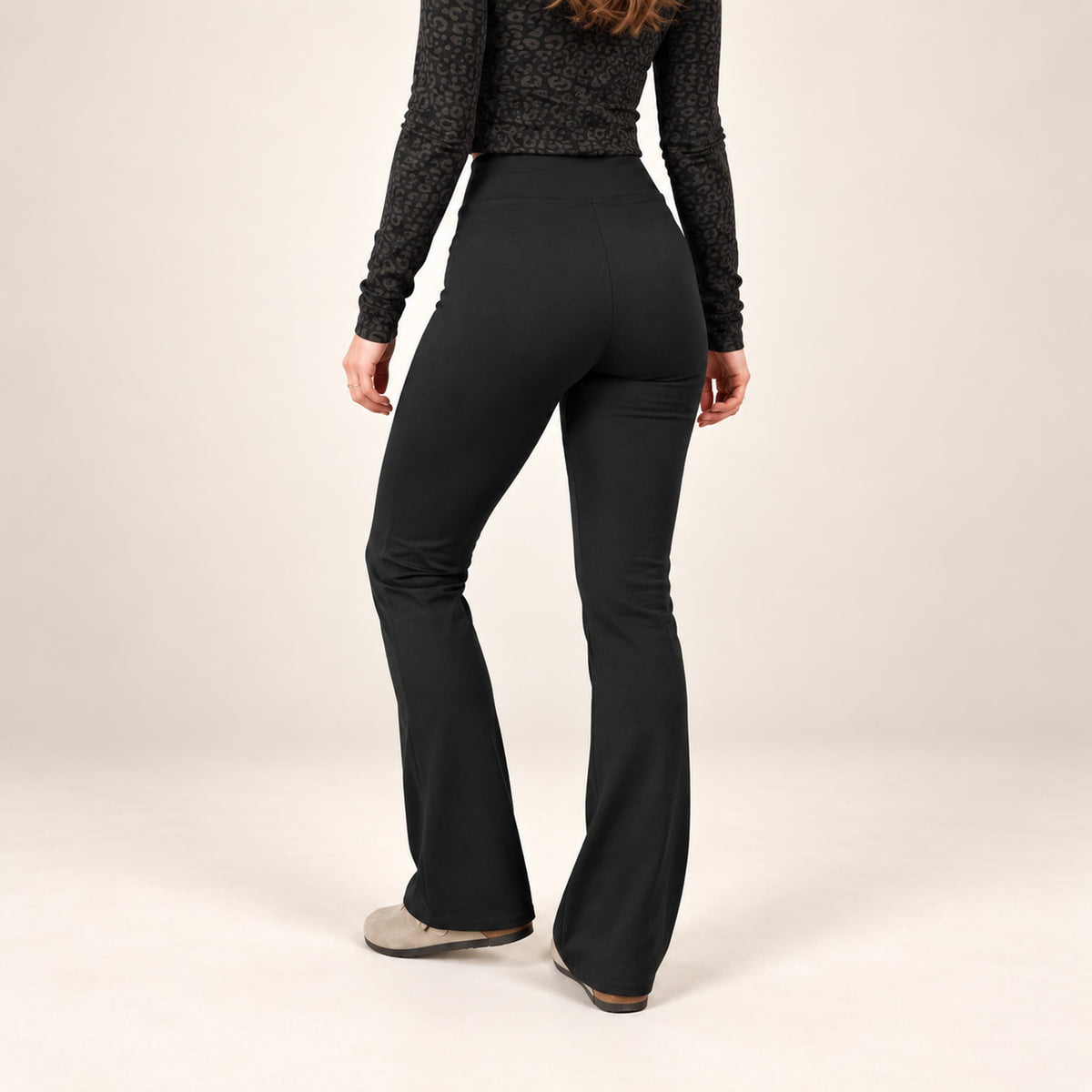A model from behind wearingk high-waisted organic leggings flared leggings, and a black patterned long-sleeve top, standing against a plain light beige studio background.