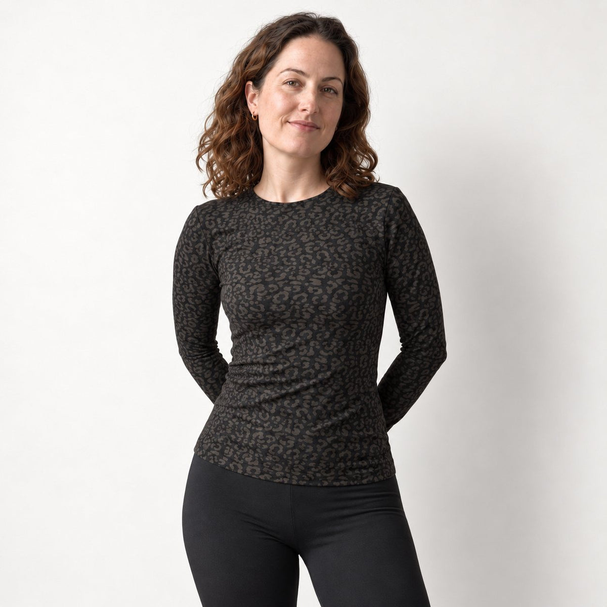 A woman modeling a black leopard long sleeve top with a dark brown and black leopard print, paired with black leggings, against a plain light gray background.