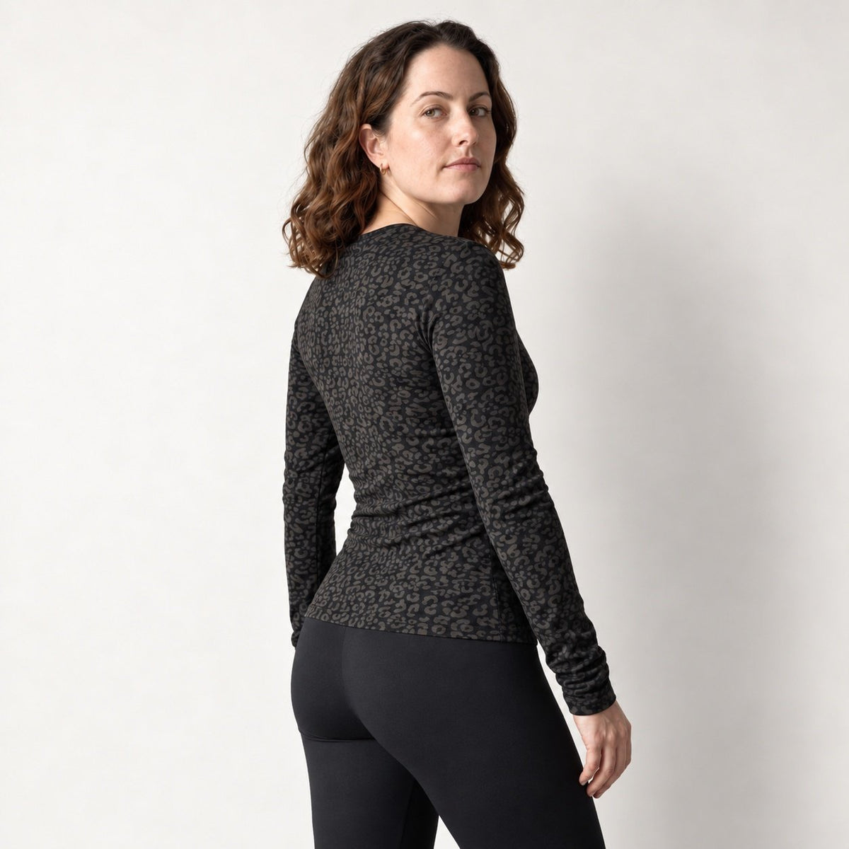 A woman wearing a fitted black leopard long sleeve top with a dark gray and black leopard print, shown from a three-quarters back view against a plain light gray studio background.