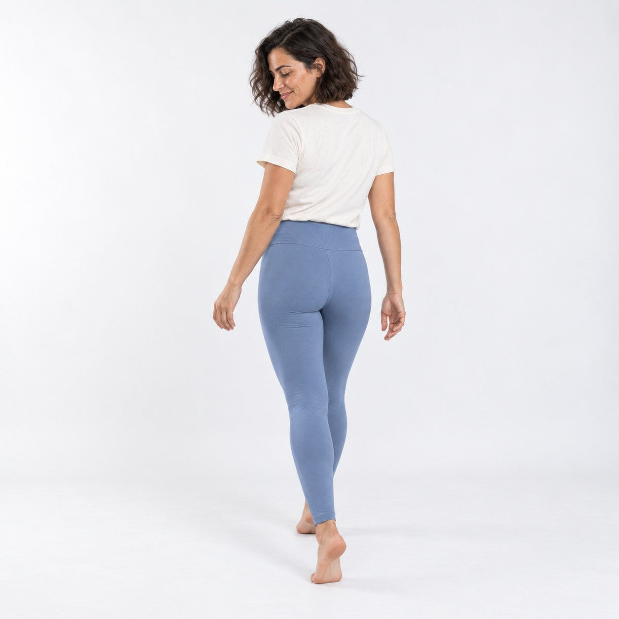 A woman wearing high-waisted, light blue  organic cotton high waist leggings  and a white t-shirt, shown from a back-three-quarter view against a plain white studio background.