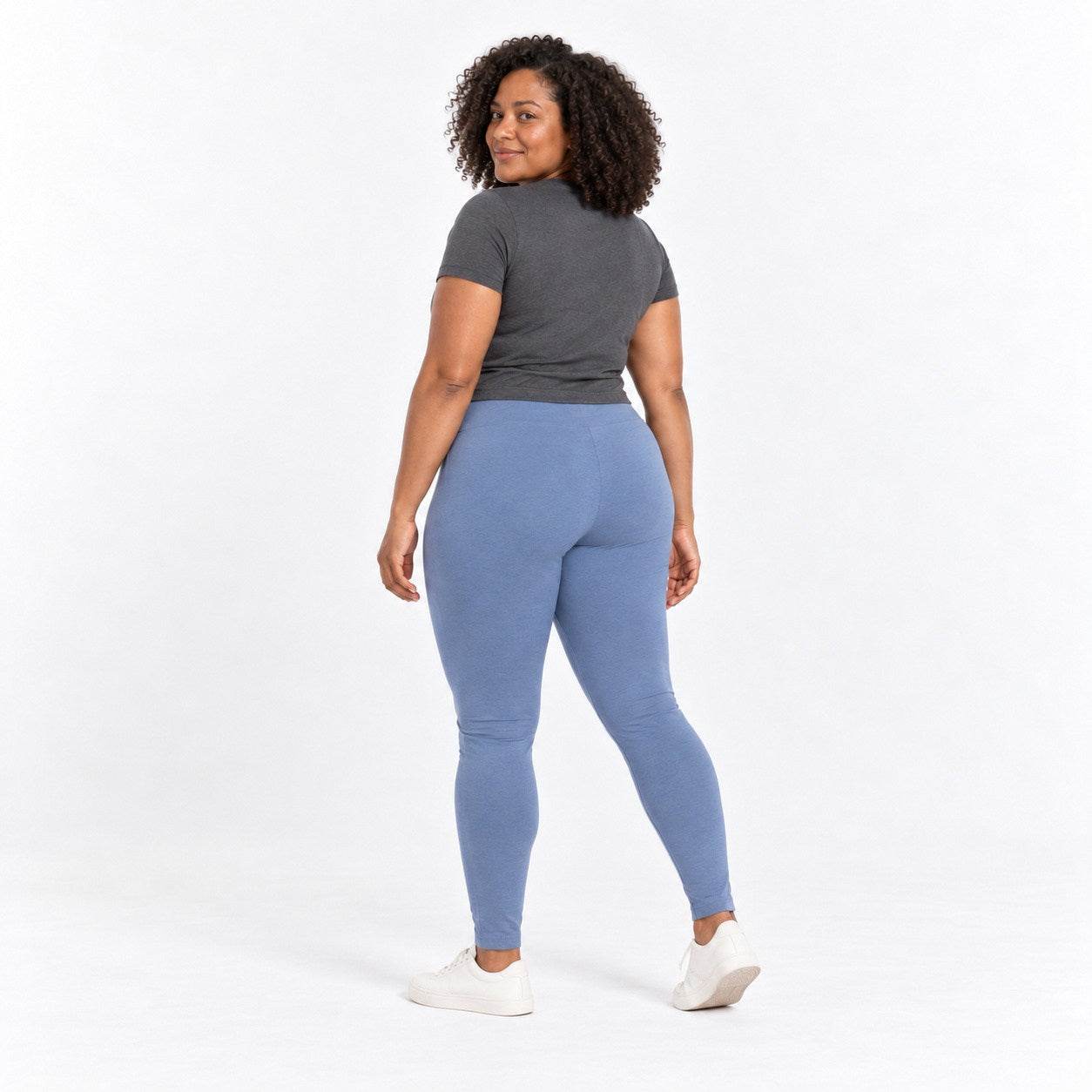 A woman is shown from the back wearing a dark gray short-sleeve t-shirt, dusty blue  organic cotton high waist leggings , and white sneakers against a plain white studio background.