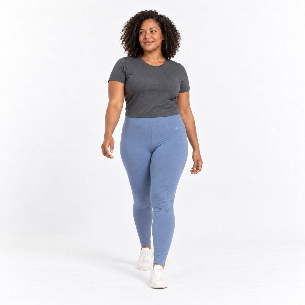 A woman modeling a dark gray short-sleeve t-shirt and light blue  organic cotton high waist leggings with white sneakers, walking against a plain white studio background.