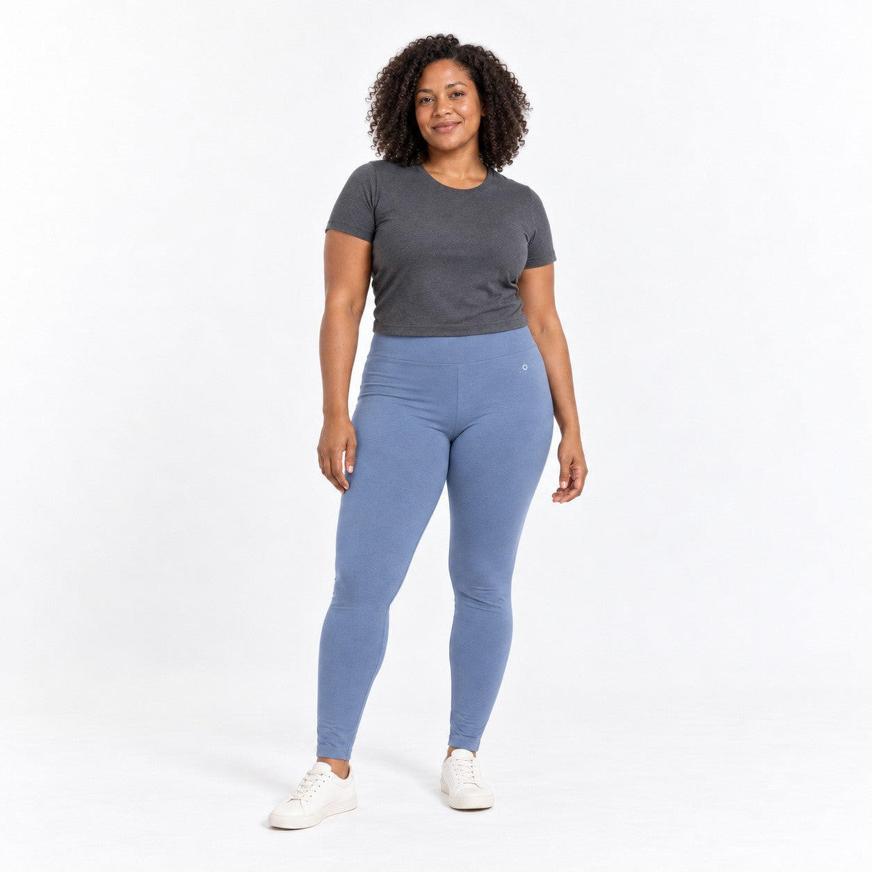 A woman modeling a dark gray short-sleeve t-shirt and light blue h organic cotton high waist leggings  with white sneakers, standing against a solid white studio background.
