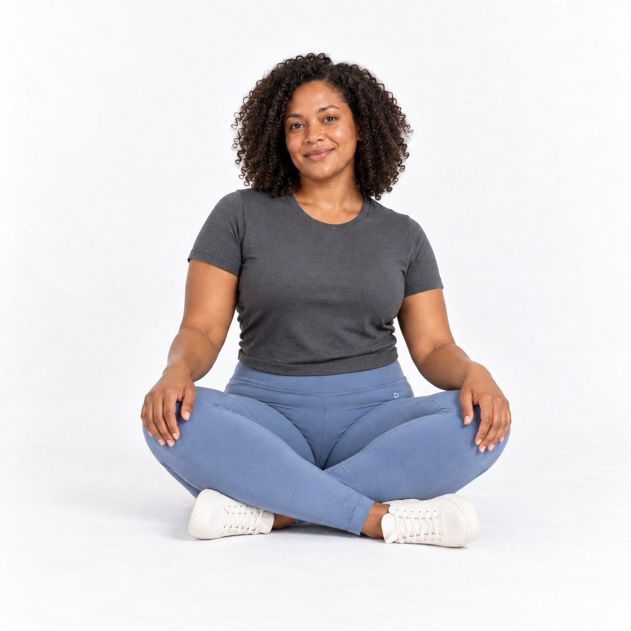 A woman with curly hair sits cross-legged on a white studio background, wearing a dark gray short-sleeve t-shirt, slate blue  organic cotton high waist leggings , and white sneakers.