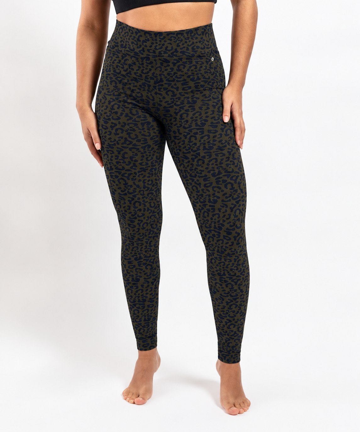 A model wearing high-waisted leopard-print leggings in dark olive green and black, standing barefoot against a plain white studio background.