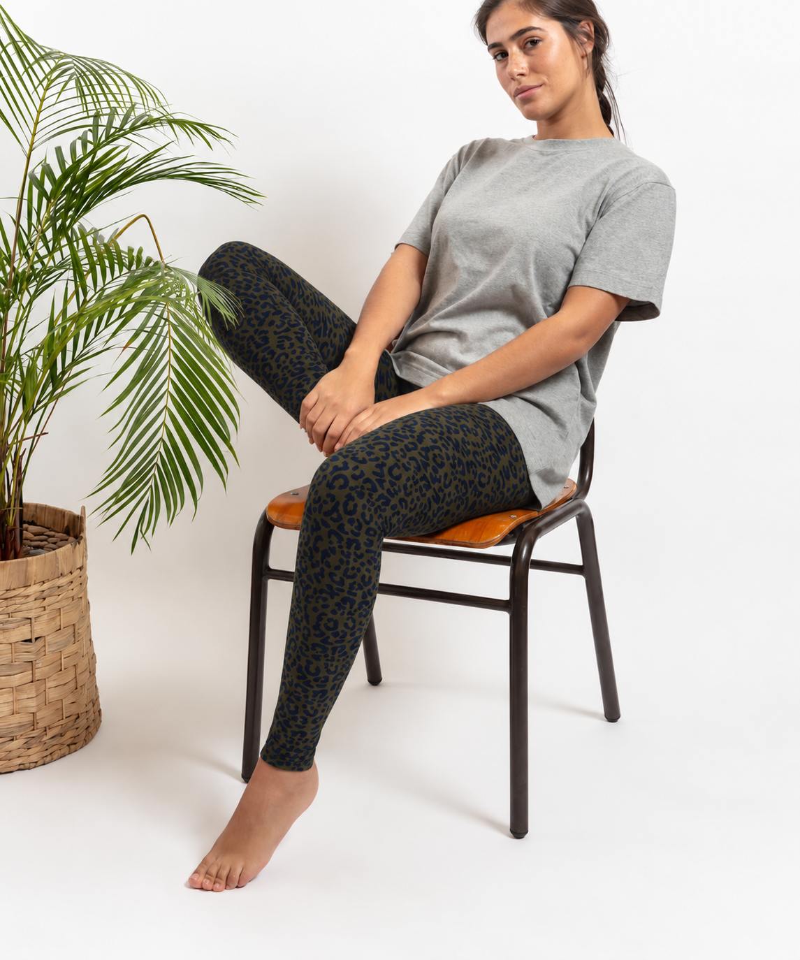 A woman sits on a black chair with a brown cushion, wearing green leopard print leggings  and a light gray t-shirt. A potted palm is on the left against a white studio background.