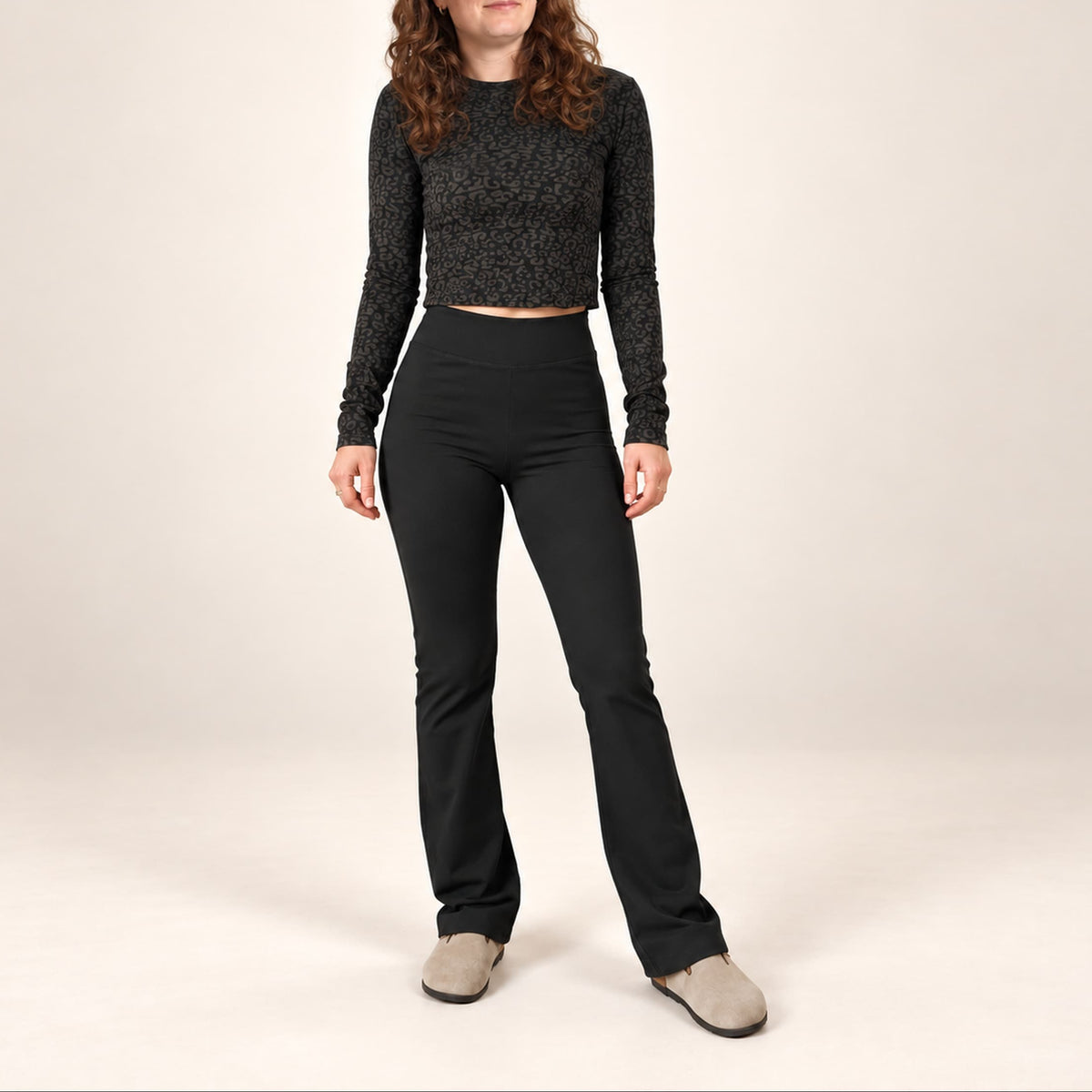 A woman modeling a black textured long-sleeve crop top and black high-waisted flared pants, paired with light gray slip-on shoes, against a plain beige studio background.