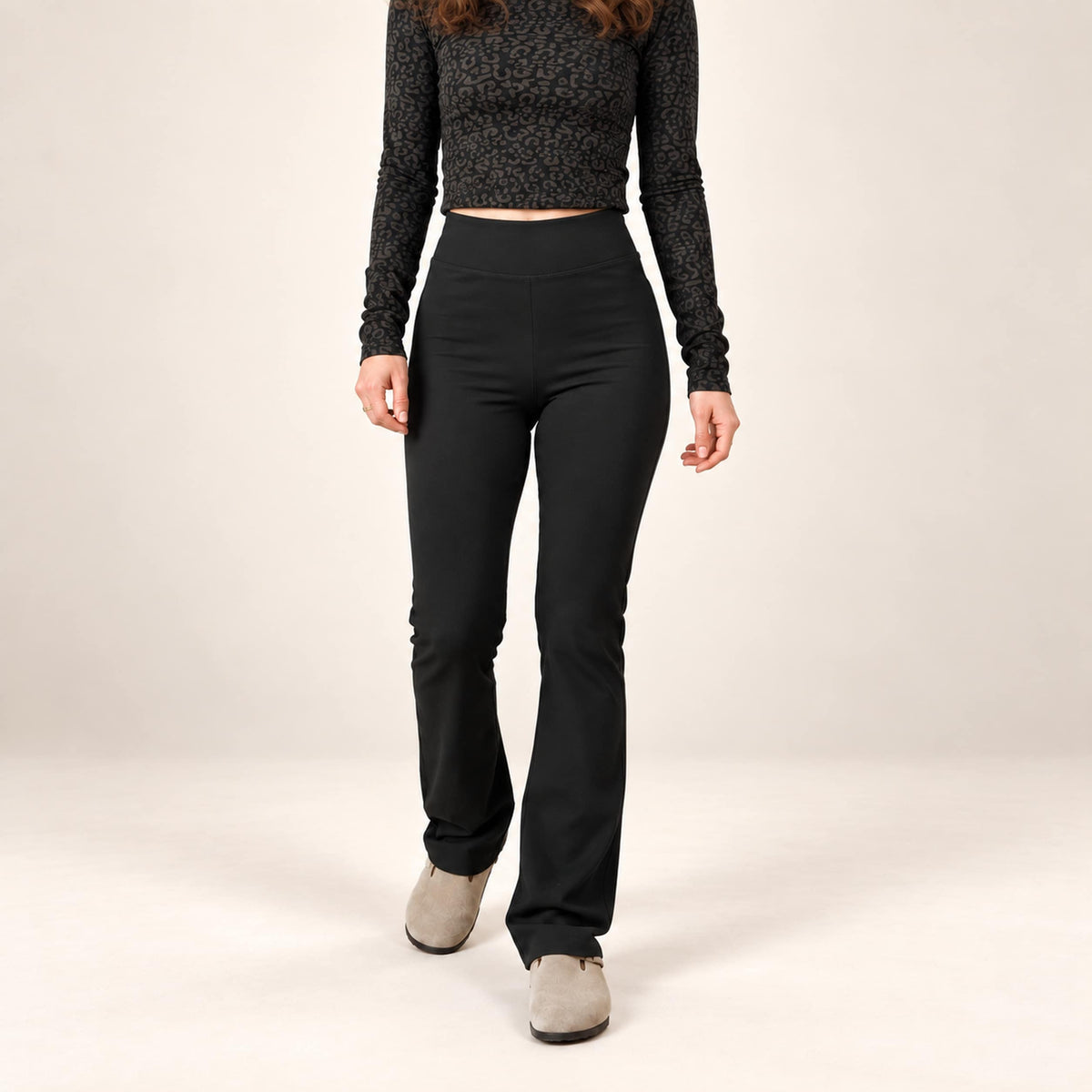 A model wearing black high-waisted organic leggings flared leggings, a black patterned long-sleeve crop top, and light gray slip-on shoes, standing against a plain light beige studio background.