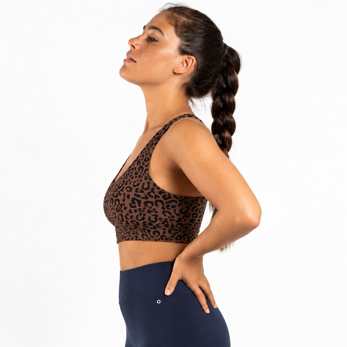 A woman in a side profile wearing a brown and black leopard print sports bra and navy blue leggings, standing against a solid white background.