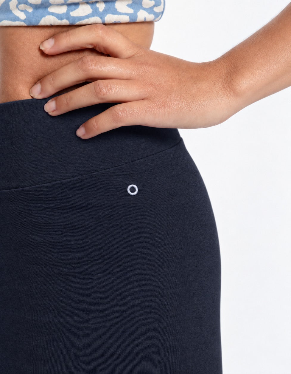Close-up of a person wearing a organic cotton high waist leggings deep blue color, showing the waistband, and a small white circular detail, with one hand resting on the hip. A patterned blue top is partially visible at the top edge against a light gray background.