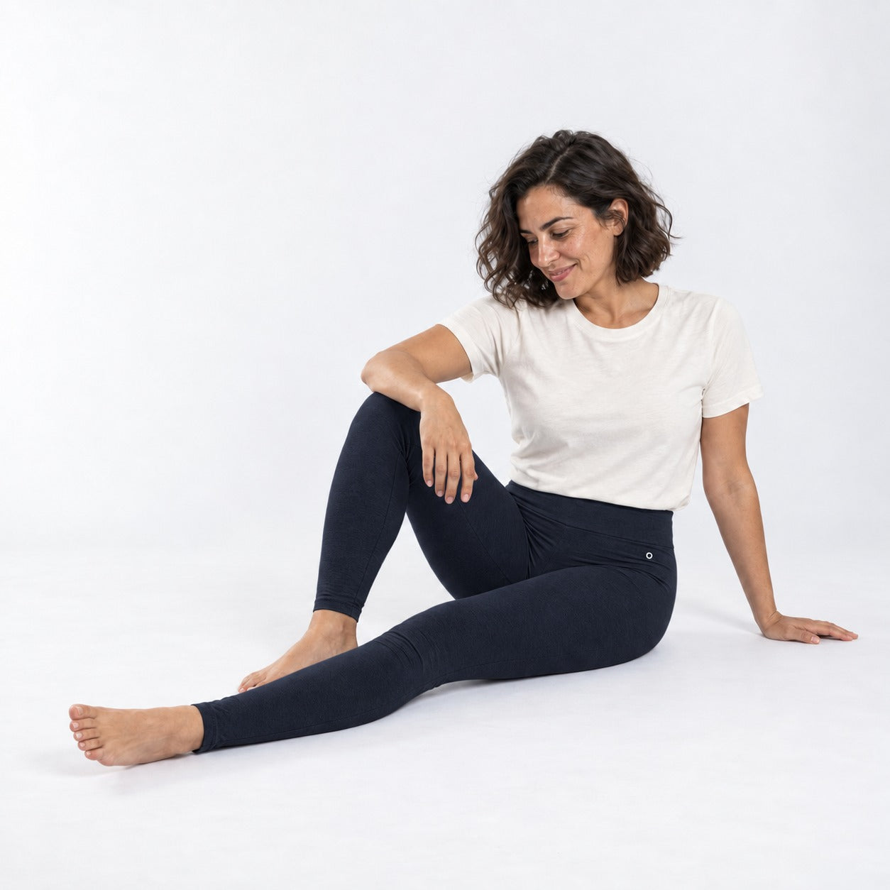A woman sitting on a white studio floor wearing organic cotton high waist leggings deep blue color and a white t-shirt.