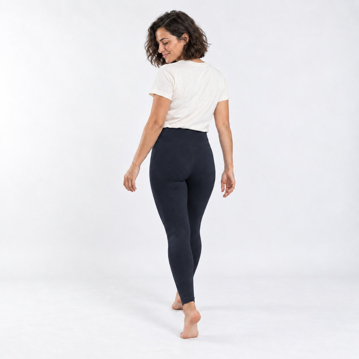 A woman is shown from behind wearing dark navy high-waisted leggings and a white t-shirt, standing barefoot against a plain white studio background.