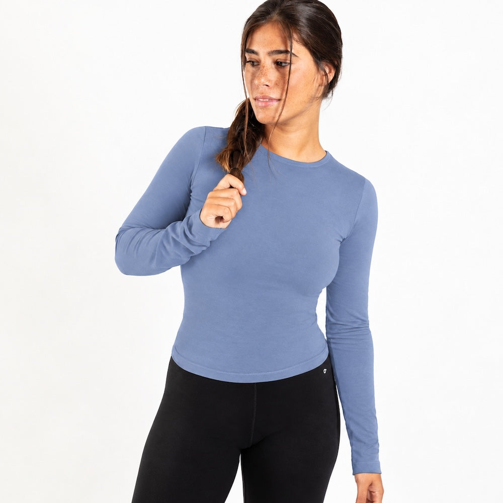 A woman modeling a fitted, organic cotton long sleeve shirt  in dusty blue, paired with black leggings, against a plain white studio background.