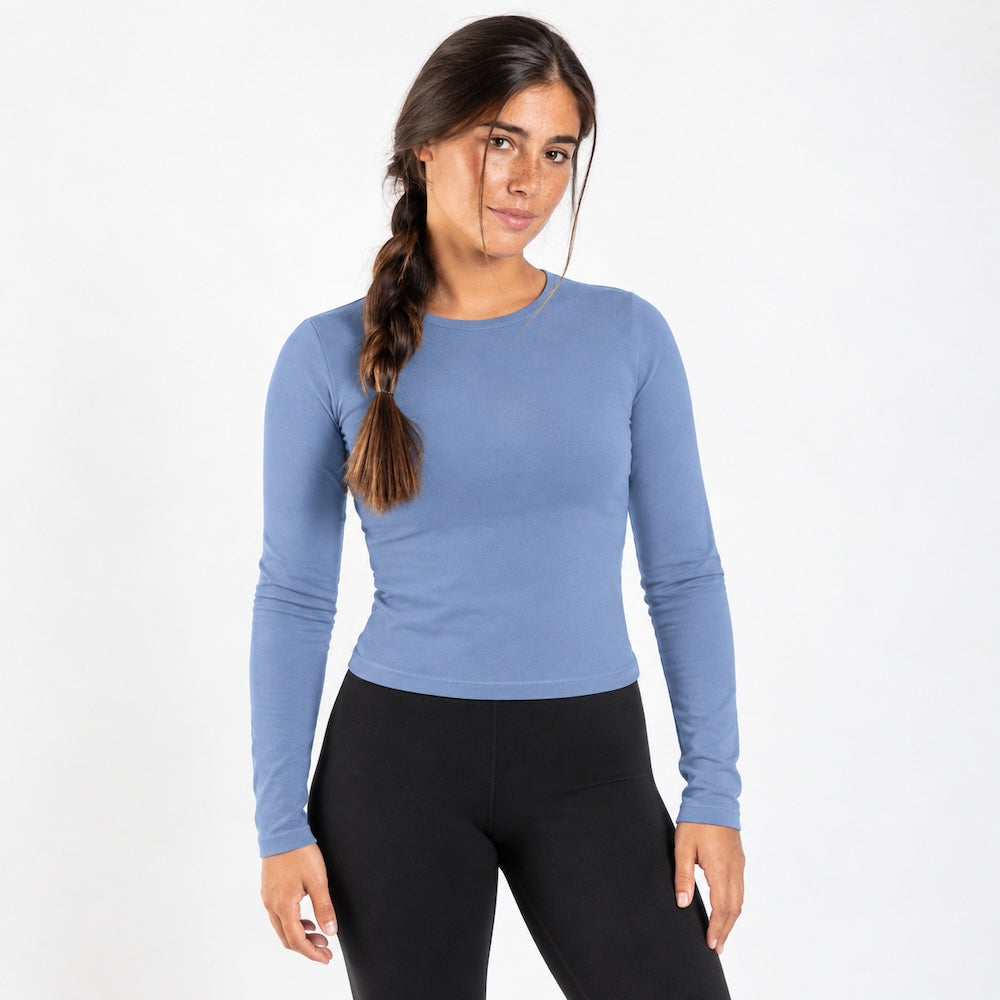 A woman modeling a fitted, organic cotton long sleeve shirt  in dusty blue, paired with black leggings, against a plain white studio background.