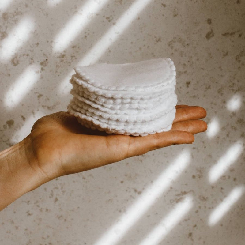 Stack of white cotton pads held in a hand against a textured beige wall.