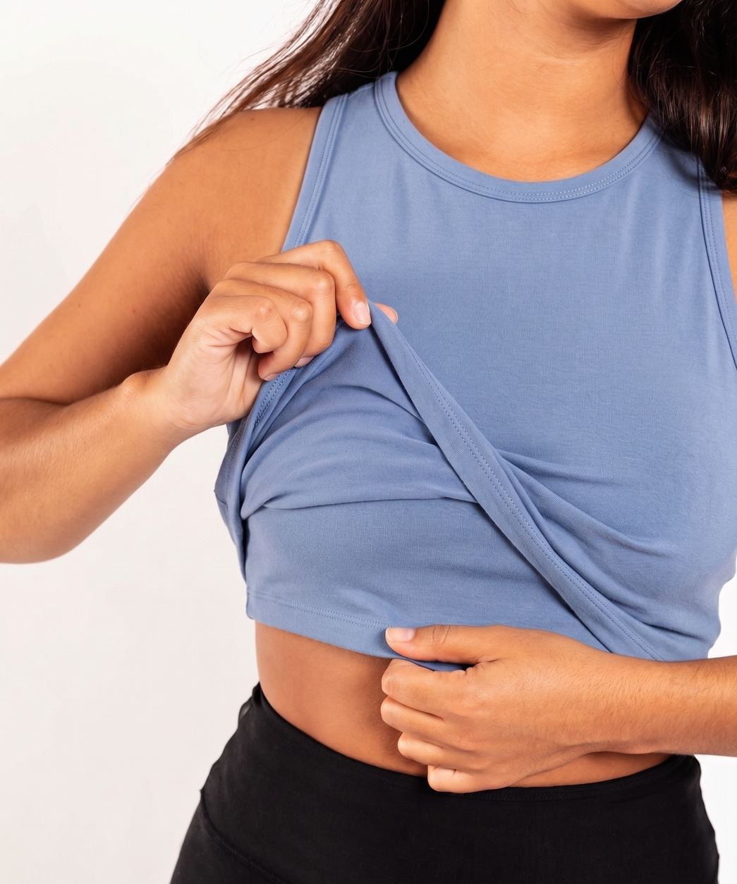 A close-up of a woman wearing a blue Organic Cotton Tank Top and black bottoms, demonstrating how to wear a crossover wrap tank top by lifting the front layer with one hand.