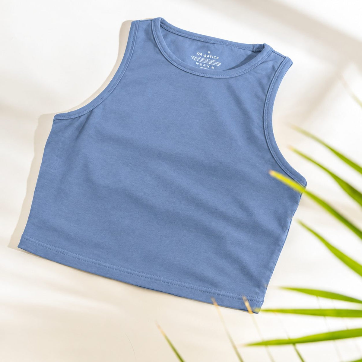 A sleeveless cropped  Organic Cotton Tank Top with built in bra  in dusty medium blue, laid flat on a light cream background with soft shadows and green plant leaves at the edge.