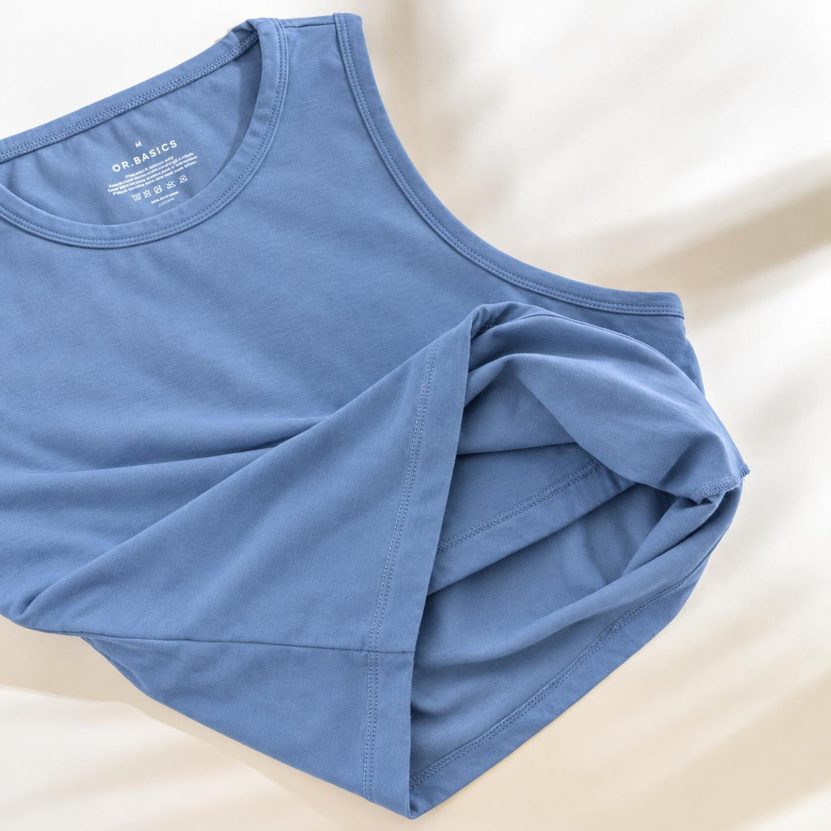 A folded light blue Organic Cotton Tank Top with built in bra  with a crew neck, laid flat on a soft beige fabric background. The brand name 'ORIGAMICS' is visible on the inner neck label.